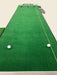 Green golf practice mat with two golf balls on a beige floor.
