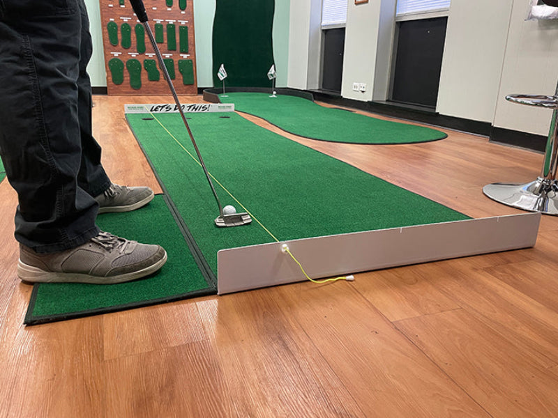 Person practicing golf on a simulator with green mat and wooden floor.