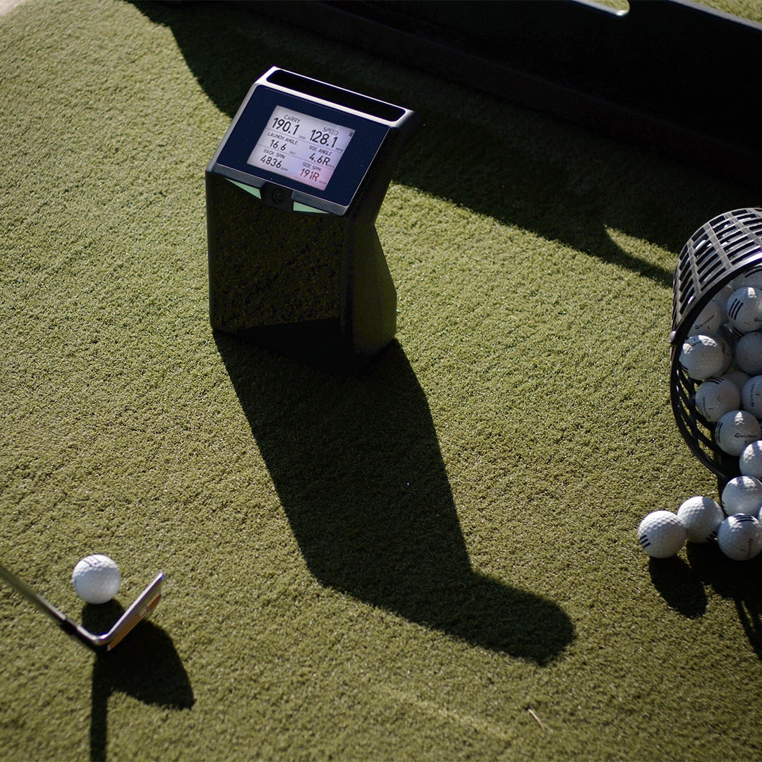 Golf ball on a tee with TruGolf Launch Box device and basket of golf balls on a green surface