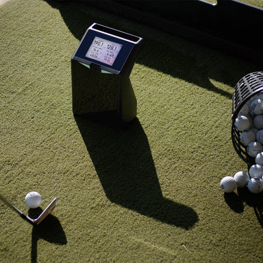 Golf ball on a tee with TruGolf Launch Box device and basket of golf balls on a green surface