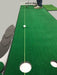 Putting green setup with a green mat, white ball, and hole on a gray floor.