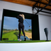 Person playing golf with simulator and a screen displaying a golf course.
