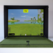 Golf simulator setup with screen displaying a golf course and a green mat on the floor.