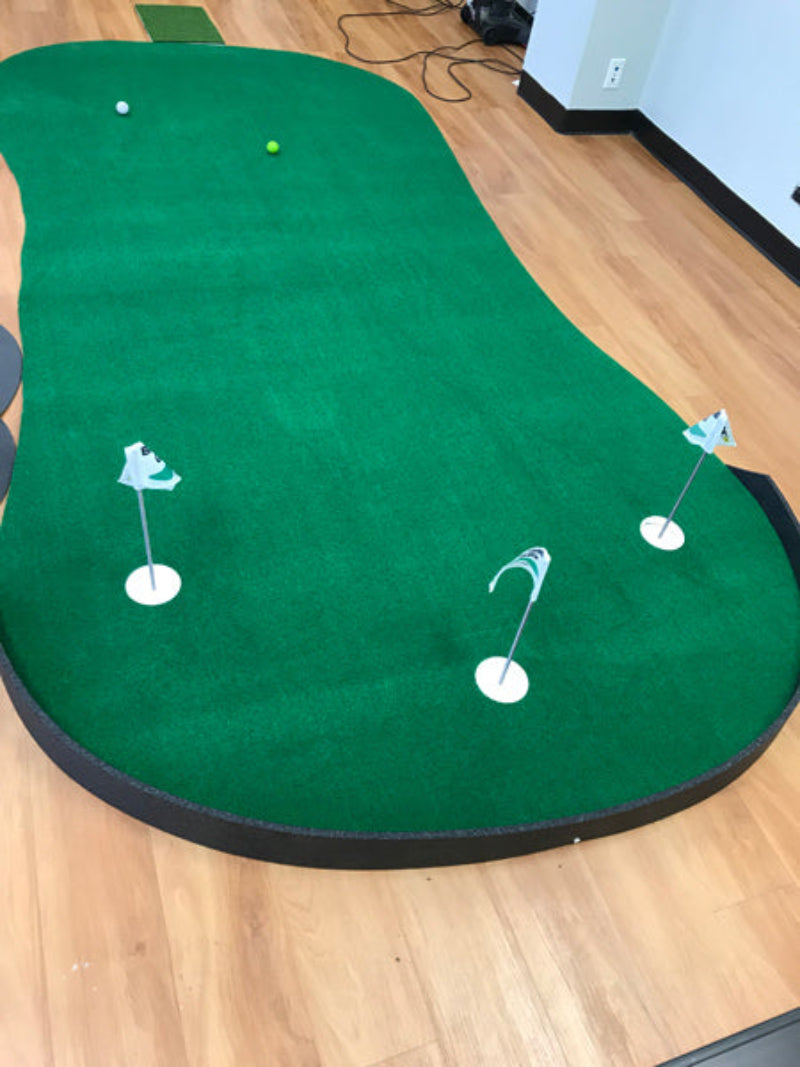 Green indoor putting green with white cups and a ball on a wooden floor.