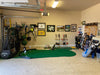 Golf practice area with green mat, clubs, and equipment in a garage setting.