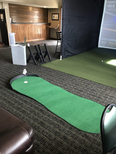 Indoor putting green with simulator screen and club holder in a room.