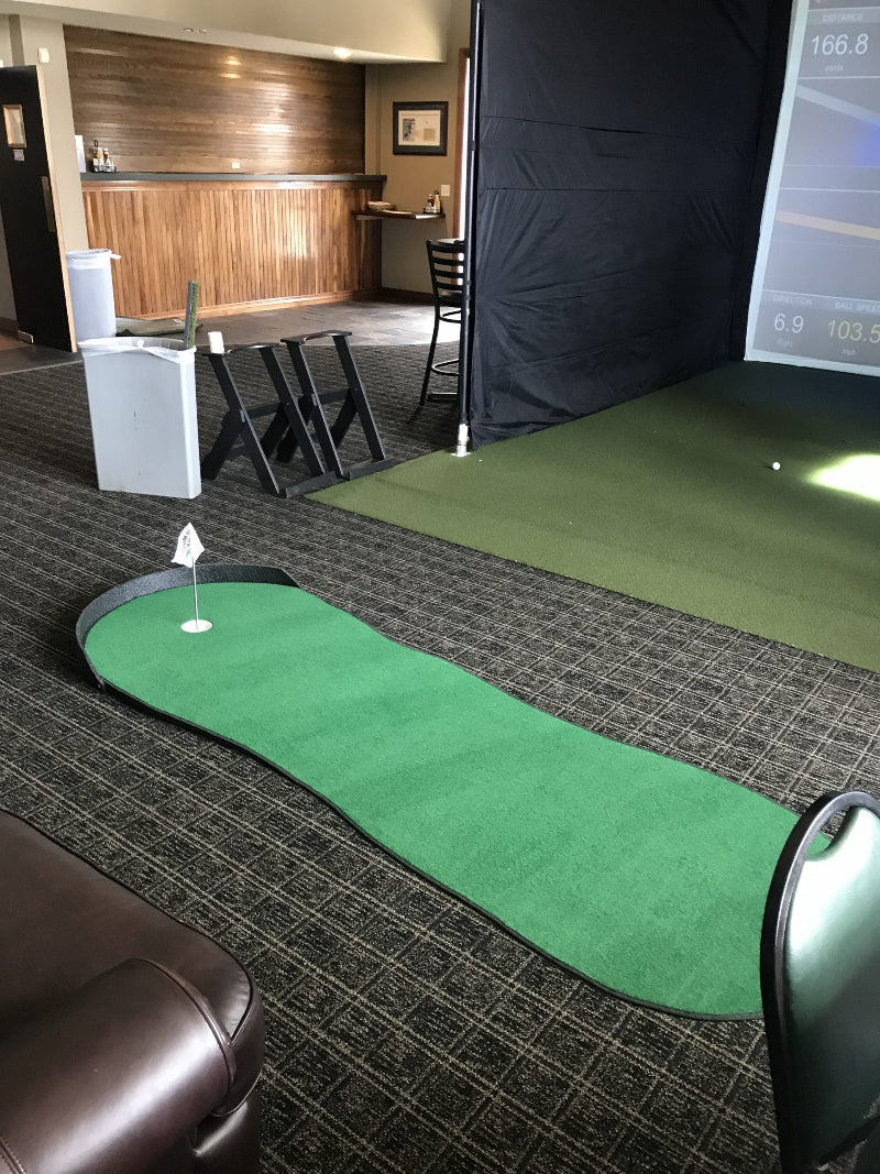 Indoor putting green with simulator screen and club holder in a room.