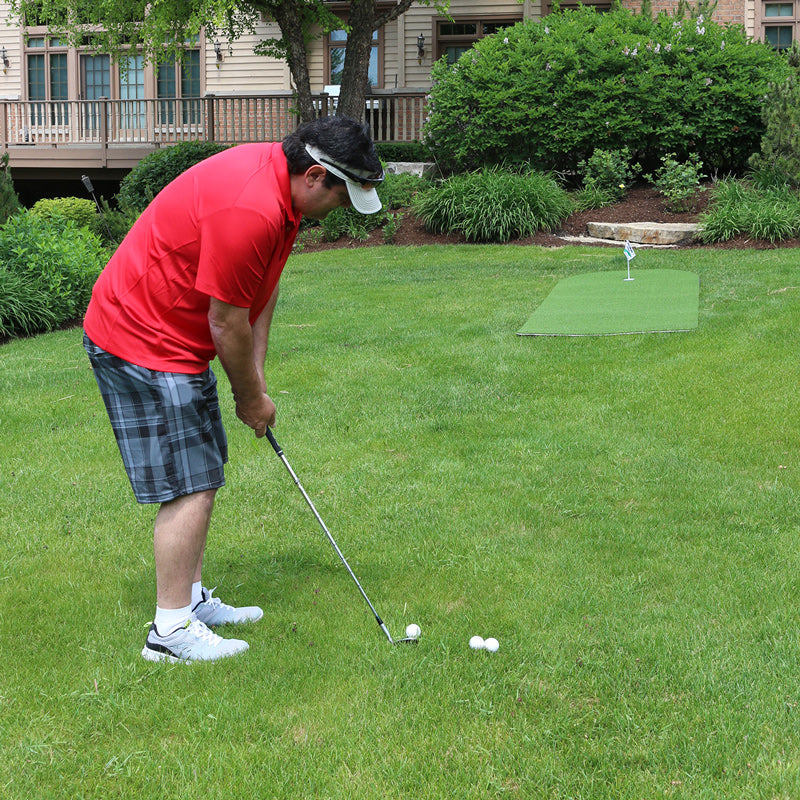 Man in red shirt and plaid shorts playing golf on a green lawn with a house in the background.